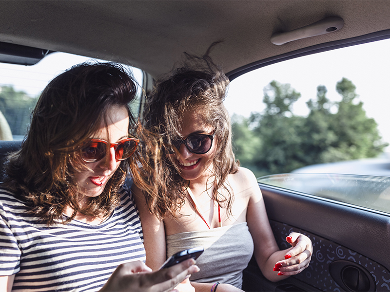 Smiling young women sitting in car and texting with smartphone