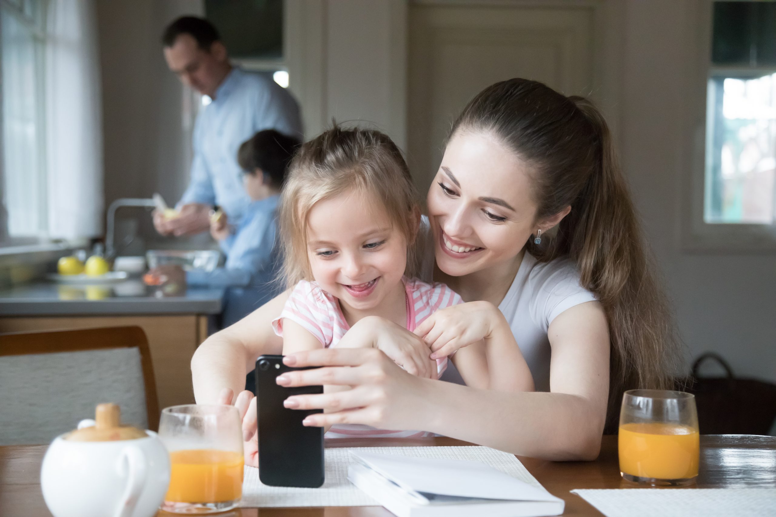 Happy mother and daughter sitting at kitchen table using smartphone