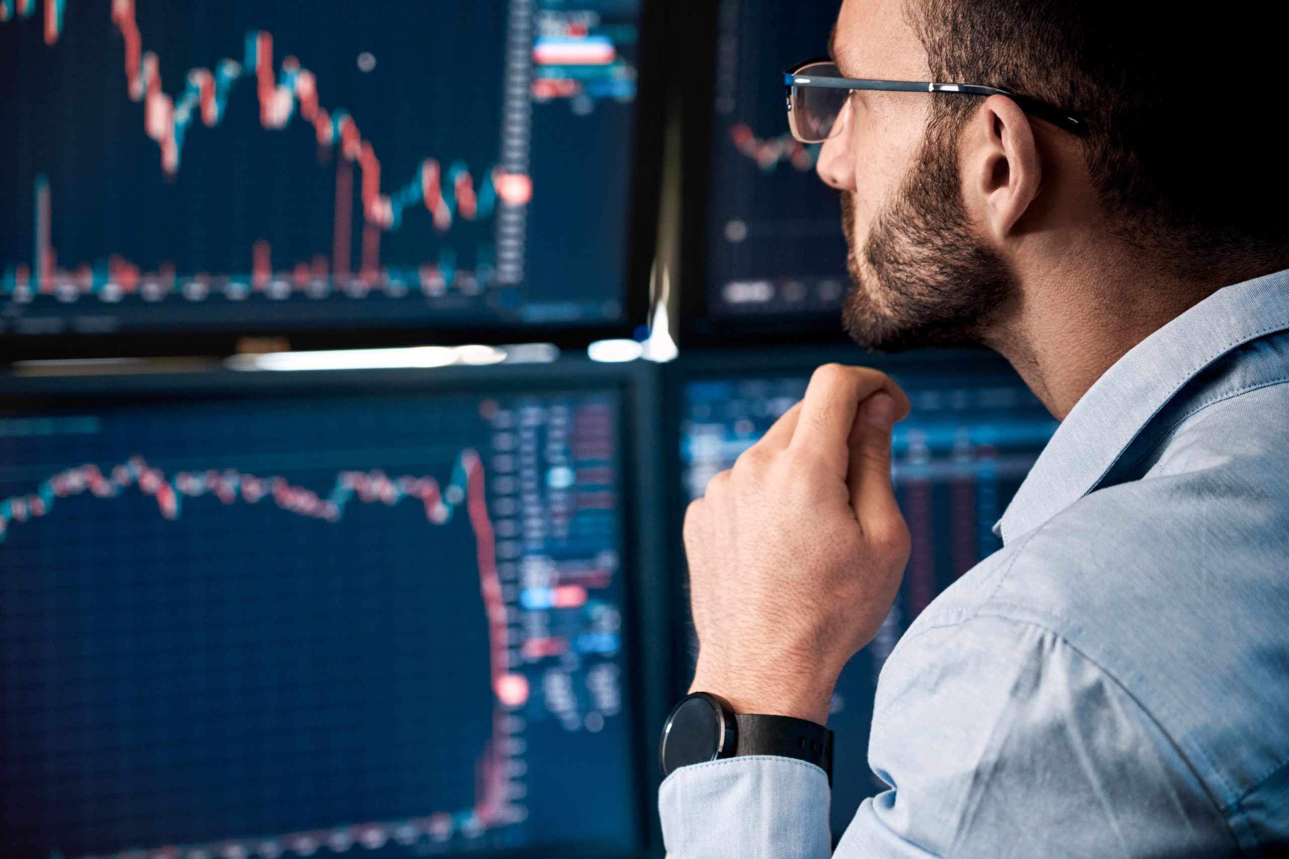 Closeup of bearded man with glasses sitting in front of four monitors